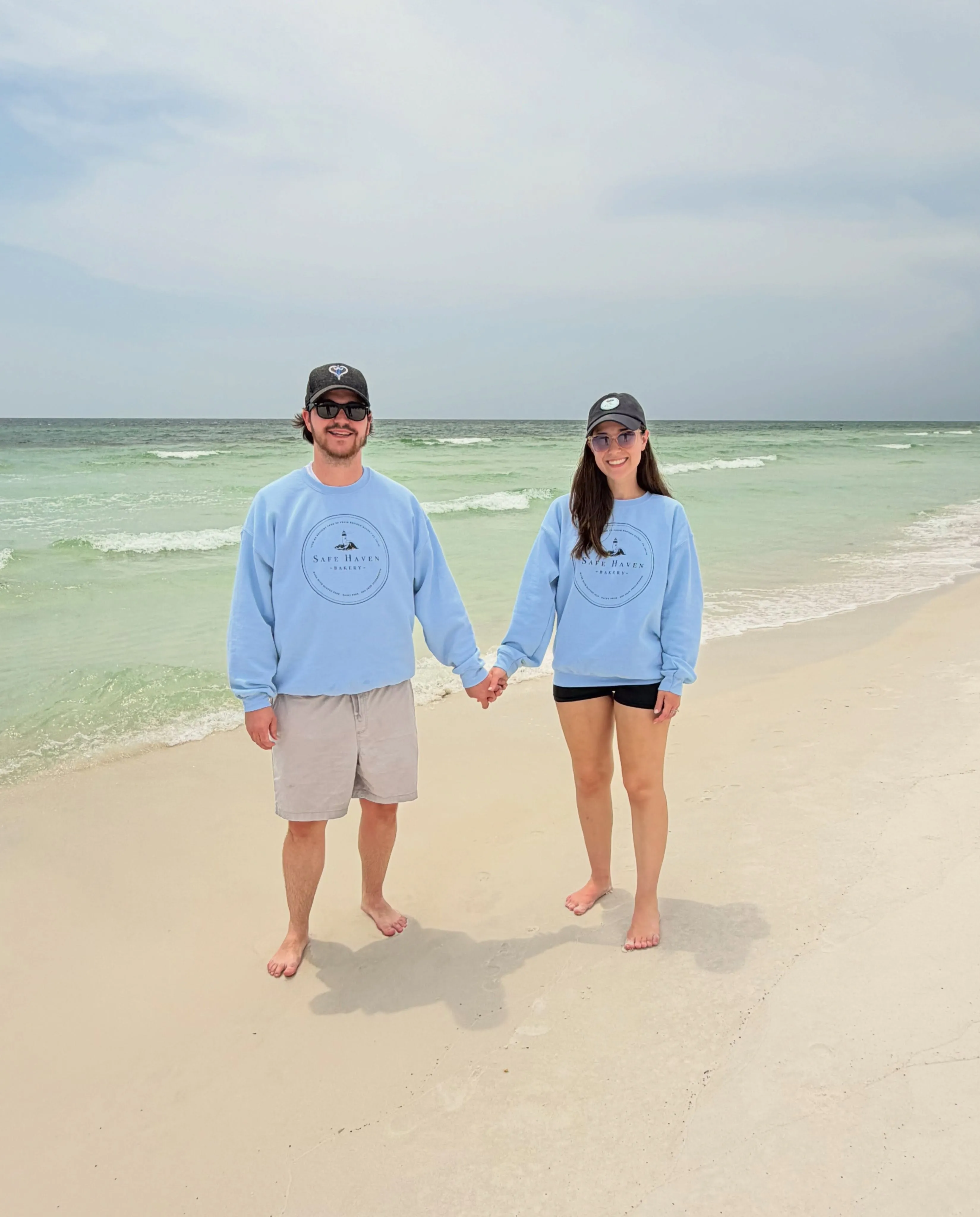 Sophie and Hunter holding hands on the beach in matching Safe Haven sweatshirts