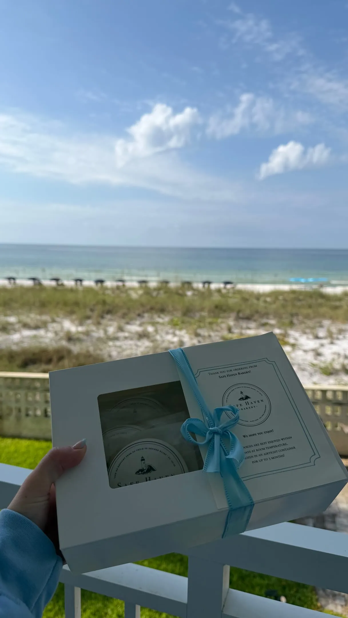 A white Safe Haven gift box tied with a light blue ribbon, held up on a balcony above the beach